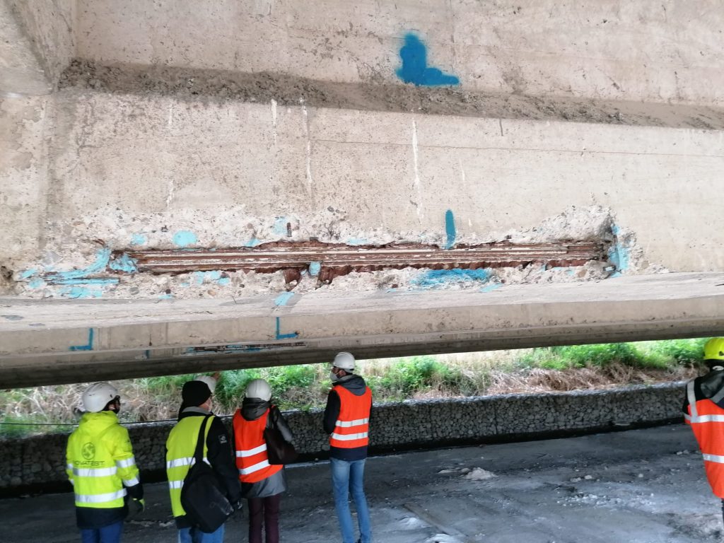 Ispettori in formazione osservano un impalcato da ponte con degrado del calcestruzzo e armature esposte durante una sessione pratica di esame visivo